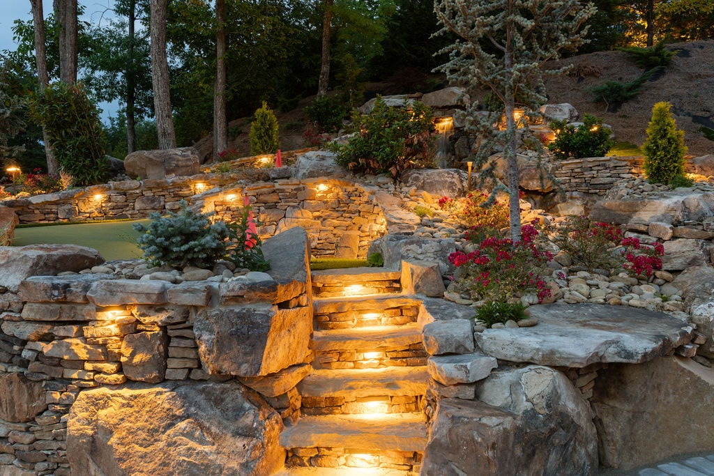 Lit stone staircase winding through the Excalibur putt-putt course at night with flowering landscaping